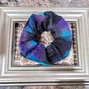 May include: A galaxy print scrunchie with a dark blue, purple, and turquoise color scheme. The scrunchie is made of fabric and has a white polka dot pattern.