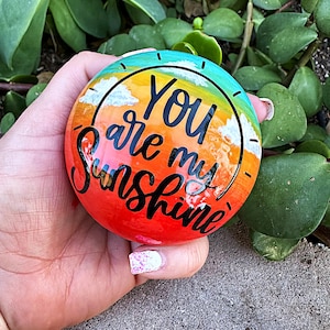 Op de afbeelding: Een beschilderde steen met een zonsondergang achtergrond en de woorden "You are my Sunshine" in zwarte letters.