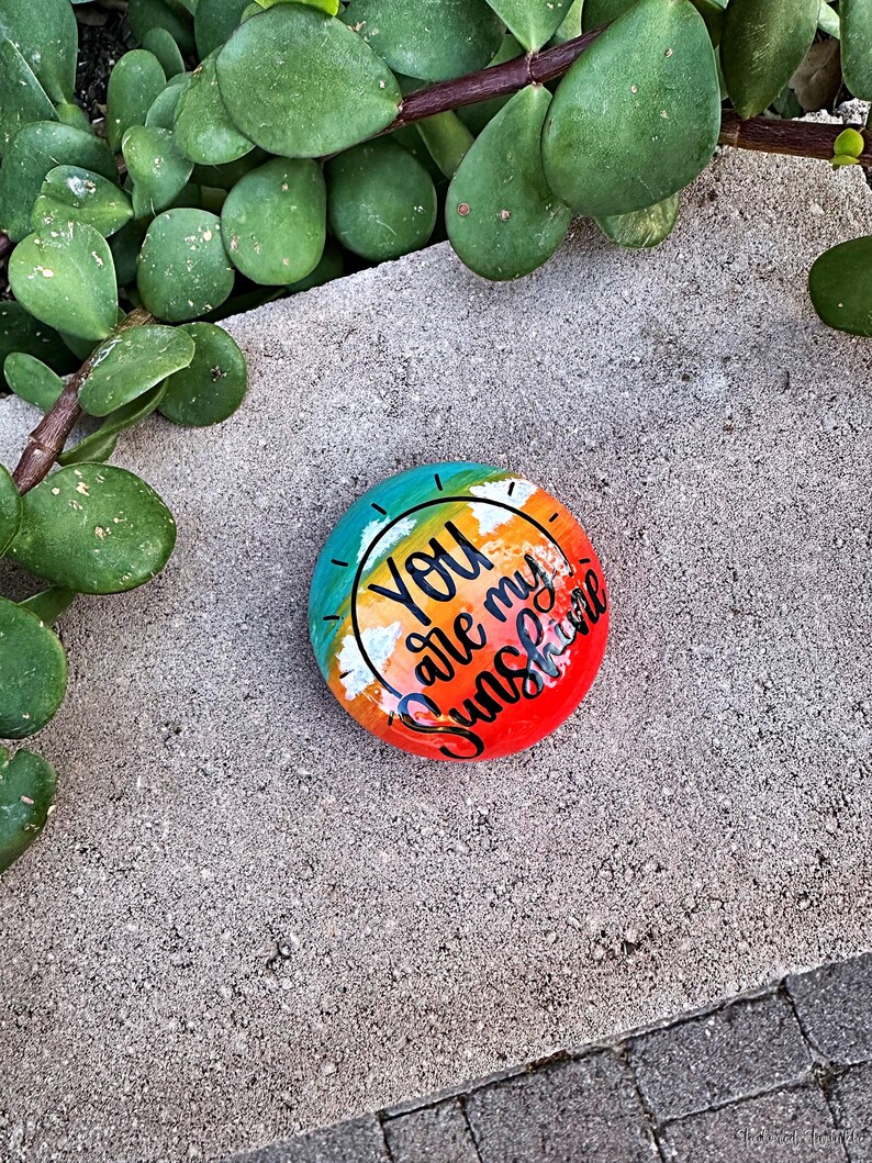Op de afbeelding: Een beschilderde steen met een regenboogachtergrond en de woorden "You are my sunshine" in zwarte letters.