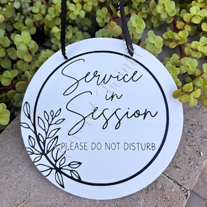 以下が含まれることがあります： 「Service in Session」と「PLEASE DO NOT DISTURB」という黒い文字が入った白い丸いサイン。サインには黒い縁と黒い花の模様があります。吊り下げ用の黒いリボンが付いています。