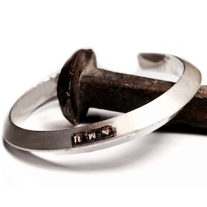 May include: A silver bracelet with a hammered finish and a small engraved inscription that reads "N.M.W.". The bracelet is resting on a rusty nail.