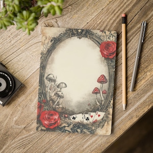 May include: A decorative notebook with an oval frame design, featuring mushrooms, roses, and playing cards. The notebook is on a wooden surface, with a pencil and pen nearby. The design has a vintage, fairytale aesthetic.