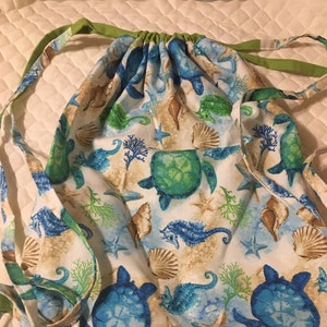 May include: Drawstring backpack featuring a sea life pattern. The fabric displays blue turtles, seahorses, starfish, and seashells on a white background with blue and tan accents. The drawstring and straps are green.