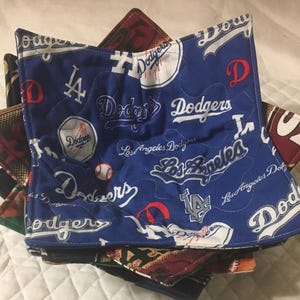 May include: A stack of quilted fabric bowl cozies. The top cozy is blue with white text and logos for the Los Angeles Dodgers baseball team. Other cozies in the stack have different patterns and colors.