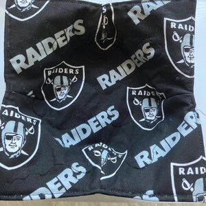 May include: Black and white Raiders bowl cozy. The cozy features the Raiders logo and the word "Raiders" in white. The bowl cozy is quilted and has a square shape. The background is white.