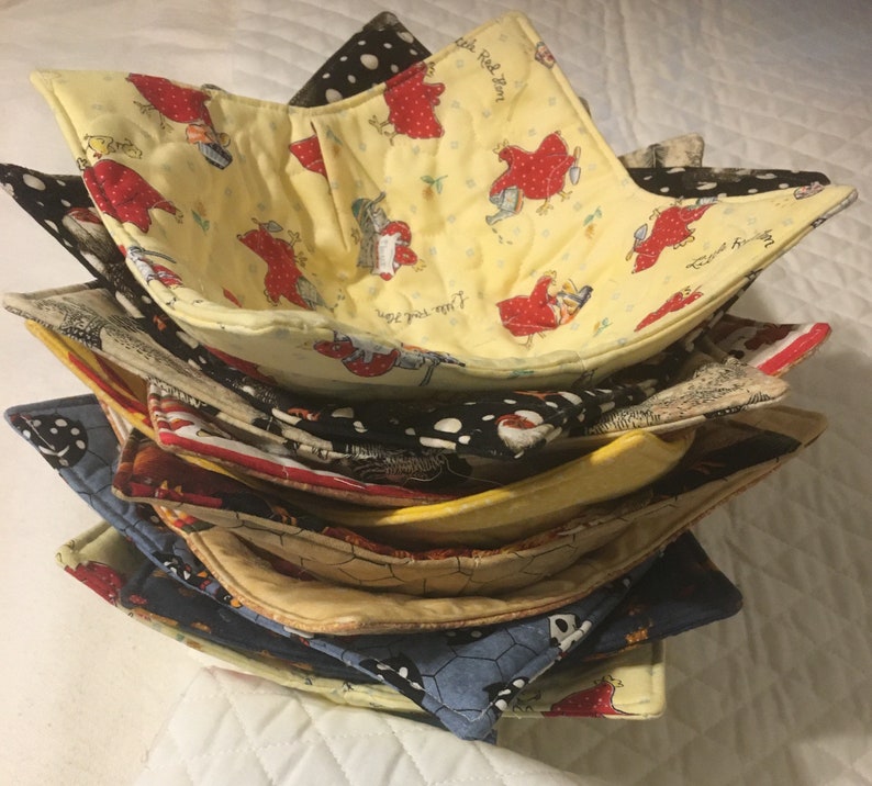May include: A stack of handmade fabric bowl cozies in various patterns. The top cozy is yellow with a red chicken print. Other cozies feature polka dots, patriotic designs, and other colorful patterns. These cozies are designed to hold bowls and protect hands from heat.