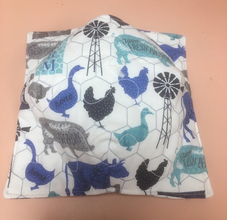 May include: A square, white bowl cozy with a farm animal print. The print includes blue, gray, and black chickens, cows, and geese, along with windmills and the words "Farm Fresh Everyday". The cozy is designed to hold a bowl.