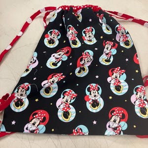 May include: Black drawstring bag featuring a repeating pattern of Minnie Mouse in a red polka dot bow and dress. The bag has red and white polka dot drawstrings and a red trim. The background is black with light blue, pink, and yellow accents.