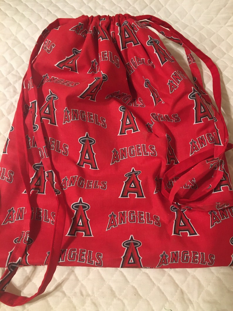 Puede incluir: Bolsa roja con cord&oacute;n con un patr&oacute;n repetido del logotipo de Los Angeles Angels y la palabra "ANGELS" en blanco. La bolsa tiene cordones rojos para cerrar.