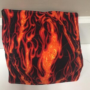 May include: A square, quilted bowl cozy featuring a vibrant flame design in shades of red, orange, and yellow against a black background. The fabric is stitched with a diamond pattern, and the edges are finished with white stitching.