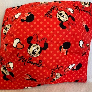May include: Red fabric bowl cozy with a polka dot pattern and Minnie Mouse illustrations. The design includes Minnie's face, bows, and the name "Minnie" in cursive. The cozy is square-shaped and appears to be quilted.