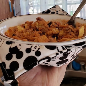 May include: A white bowl filled with a casserole dish, held by a black and white Mickey Mouse print bowl cozy. A silver spoon rests in the food. The cozy has a black border and is held by a hand.