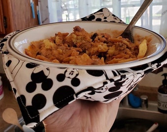 May include: A white bowl filled with a casserole dish, held by a black and white Mickey Mouse print bowl cozy. A silver spoon rests in the food. The cozy has a black border and is held by a hand.