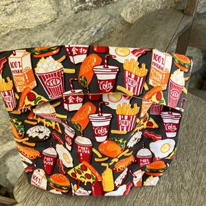 May include: A fabric pouch with a dark gray background, covered in colorful illustrations of fast food items. The design includes images of burgers, pizza, fries, cola cups, and popcorn. The pouch has two pockets and is sitting on a wooden surface.