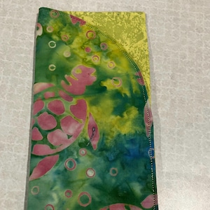 May include: A handmade fabric wallet featuring a sea turtle design. The wallet has a green, blue, and yellow batik pattern with pink sea turtles. The edges are stitched with pink thread. The wallet is rectangular and folded.