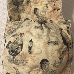 May include: Cream-colored quilted bowl cozy with a rooster and feather print. The fabric features black and white illustrations of roosters and feathers on a beige background. The cozy is designed to hold a bowl.
