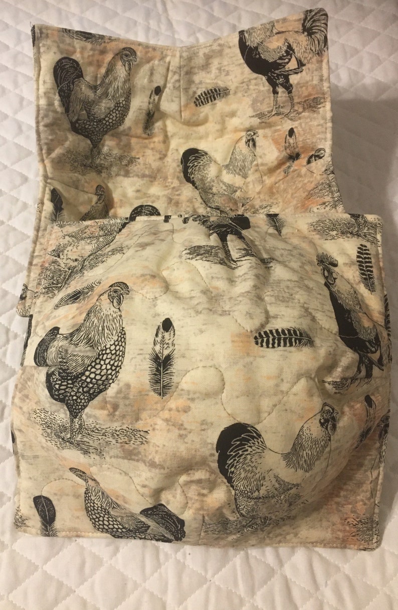 May include: Cream-colored quilted bowl cozy with a rooster and feather print. The fabric features black and white illustrations of roosters and feathers on a beige background. The cozy is designed to hold a bowl.