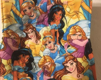 May include: A square, quilted fabric bowl cozy featuring a colorful pattern of cartoon princesses in various poses. The princesses are depicted in yellow, blue, purple, and pink dresses. The edges of the cozy are stitched with white thread.