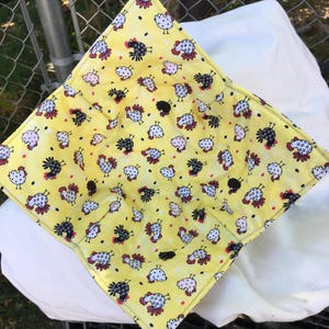 May include: A yellow fabric bowl cozy with a repeating pattern of cartoon chickens in white, black, and pink. The cozy is square with folded corners, and the background is a bright yellow color.