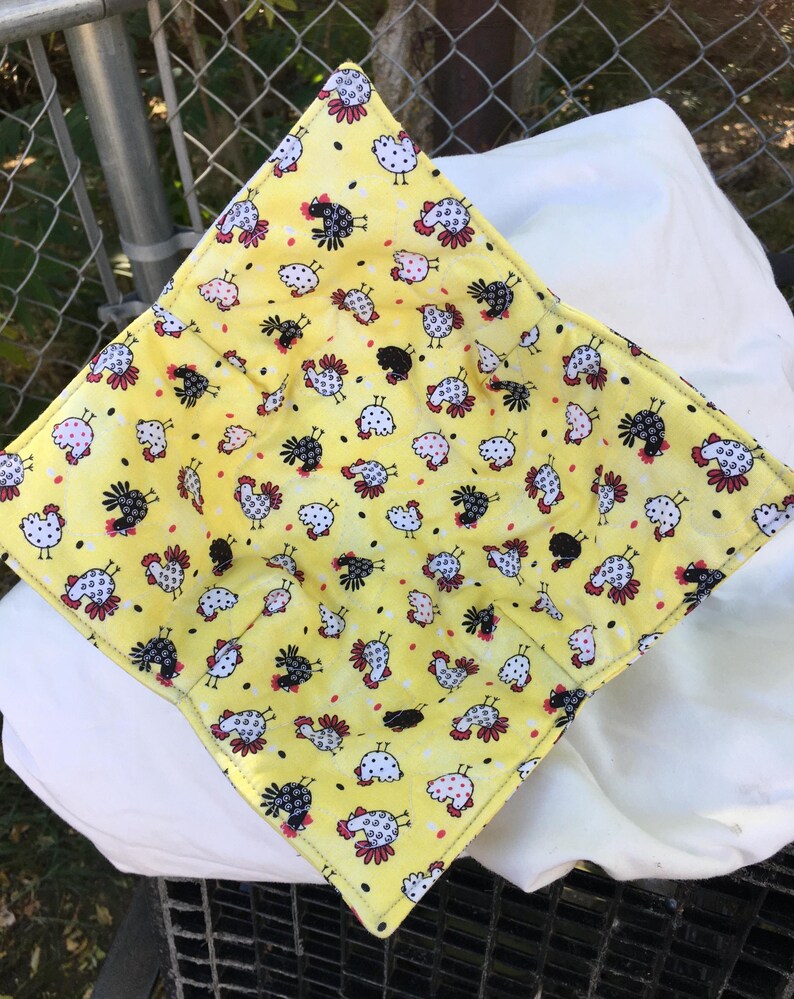 May include: A yellow fabric bowl cozy with a repeating pattern of cartoon chickens in white, black, and pink. The cozy is square with folded corners, and the background is a bright yellow color.