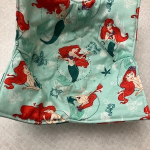 May include: A light blue fabric bowl cozy featuring a repeating pattern of a mermaid with red hair and a green tail. The mermaid is surrounded by sea creatures and floral designs. The cozy is quilted and has a square shape.