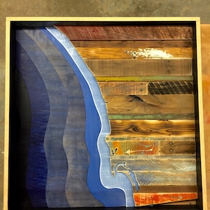 May include: A framed wall art piece made of reclaimed wood with a blue and white wave design. The wood is painted in various shades of brown, green, and red.