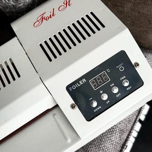 May include: A white Foil It foiler machine featuring a digital temperature display, a ready indicator light, and control buttons labelled Down, Up, On, Off, Fwd, and Rev. The device has a series of vertical ventilation slots on the top surface.