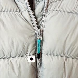 Puede incluir: Primer plano de una chaqueta acolchada gris claro con cremallera plateada. El tirador de la cremallera tiene un cordón turquesa y un extremo de plástico negro. La chaqueta tiene un diseño acolchado.