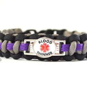 Puede incluir: Una pulsera de paracord negra, gris y morada con una etiqueta de alerta médica plateada que dice "BLOOD THINNER" con un símbolo rojo de estrella de la vida.