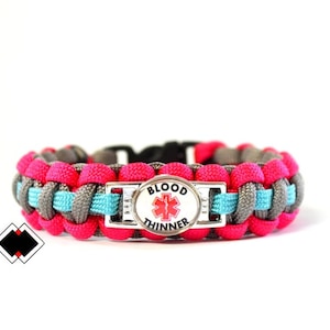 Puede incluir: Una pulsera de paracord rosa, gris y azul con una etiqueta de alerta médica plateada que dice "BLOOD THINNER".