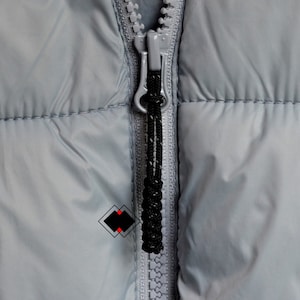 May include: Close-up of a gray zipper on a puffer jacket. The zipper has a black pull tab and is partially open.