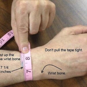 May include: A pink measuring tape is wrapped around a person's wrist to show how to measure for a bracelet. The tape measure reads 7 1/4 inches. The text says "Measure just up the arm from the wrist bone. Don't pull the tape tight. Wrist bone."