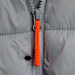 May include: Close-up of a grey zipper with an orange pull tab on a grey puffer jacket.