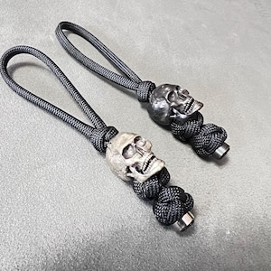 May include: Two skull-shaped zipper pulls with black cord loops. The skulls are detailed with open mouths and eye sockets. The cord is knotted below the skull and has a metal end.