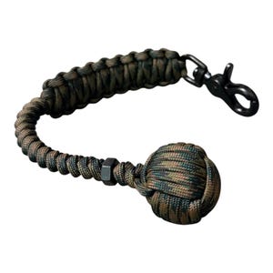May include: A keychain featuring a camouflage-patterned paracord braided into a thick strap, ending in a large spherical monkey fist knot. A black metal hex nut sits between the knot and the strap, which attaches to a black metal swivel snap hook.