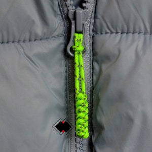 May include: A close-up of a gray zipper with a green and black rope zipper pull.