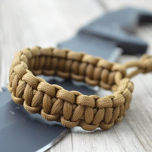 May include: A brown paracord bracelet with a woven design. The bracelet is lying on top of a black knife with a serrated edge.