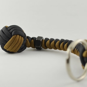 Op de afbeelding: Een zwarte en bruine paracord monkey fist sleutelhanger met een metalen moer accent. De sleutelhanger heeft een geweven bal ontwerp en een zilveren sleutelring. Het kleurenschema omvat zwart en bruin.
