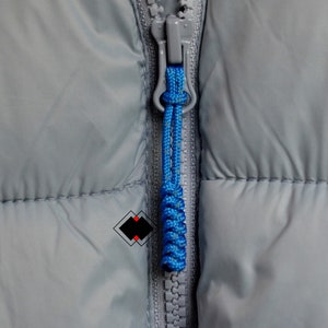 May include: Close-up of a gray zipper on a jacket with a blue cord pull. The zipper is partially closed and the cord is knotted.