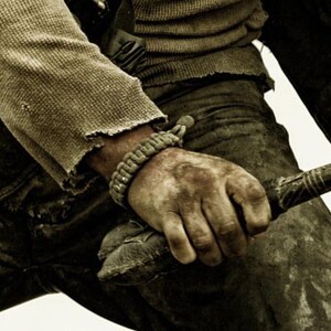 May include: A close-up of a person's hand holding a wooden club. The hand is wearing a brown paracord bracelet and is covered in dirt. The person is wearing a brown shirt and blue jeans.