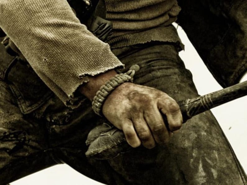 May include: A close-up of a person's hand holding a wooden club. The hand is wearing a brown paracord bracelet and is covered in dirt. The person is wearing a brown shirt and blue jeans.