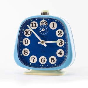 Könnte beinhalten: Ein blauer Vintage-Wecker mit hellblauem Rahmen und cremefarbenem Sockel. Das Zifferblatt ist tiefblau mit weißen Ziffern und Zeigern. Die Uhr hat einen kleinen, cremefarbenen Knopf oben.
