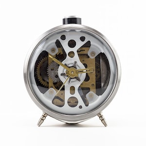 May include: A silver-coloured, round alarm clock with a transparent face showing the internal gears and mechanisms. The clock has a black top and three small, silver legs. The hands are gold-coloured.