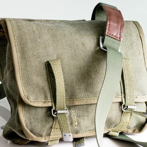 Peut inclure: Sac messager vintage en toile vert olive avec fermeture à rabat. Il est doté d'une bandoulière en cuir marron et de sangles en toile vert olive avec boucles métalliques. Le sac est de forme rectangulaire.