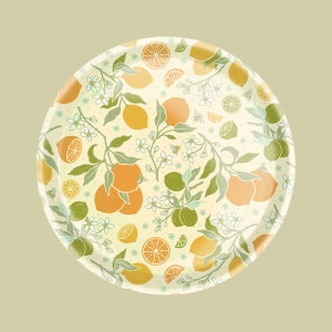May include: A round white plate with a lemon and lime pattern. The plate features lemons, limes, leaves, and white flowers. The background is a light yellow.