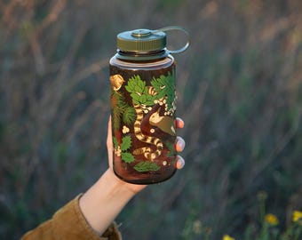 May include: A clear water bottle with a green lid and a decorative design featuring a snake, plants, and berries. The bottle is held up against a blurred background of tall grass and yellow flowers.