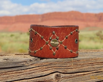 Könnte beinhalten: Ein braunes Lederarmband mit einem rautenförmigen Design, das mit weißem Garn gestickt und mit Messingnieten und einem großen Messingknopf verziert ist.