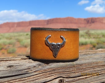 May include: A brown leather cuff bracelet with a silver bull skull design. The bracelet is on a wooden surface with a red rock background.