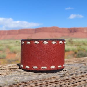 May include: A red leather cuff bracelet with white stitching. The bracelet is sitting on a wooden surface.
