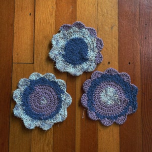 May include: Three crocheted flower-shaped pot holders in shades of purple, blue, and white. The pot holders are on a wooden floor.